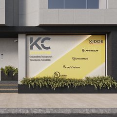 Zemin kat cephesi: siyah kaplama, sarı KC Yangın harfleri, buzlu camlı giriş kapısı ve üç basamak, logo ve partner markalarıyla grafik kaplı vitrin, önünde bitki kasaları; kaldırımda yürüyen bir kişi